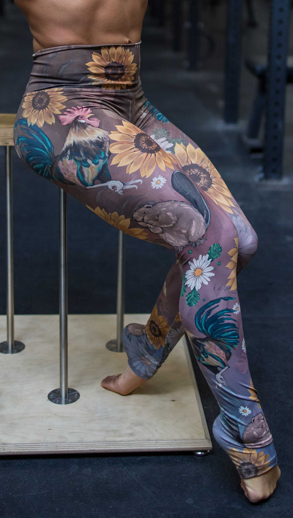 Model wearing WERKSHOP "Cocks n Beavers" Leggings. The leggings show hand painted artwork of roosters and beavers with daisies and sunflowers over an ombreback ground that goes from a warm brown on top to slate blue at the leg opening.