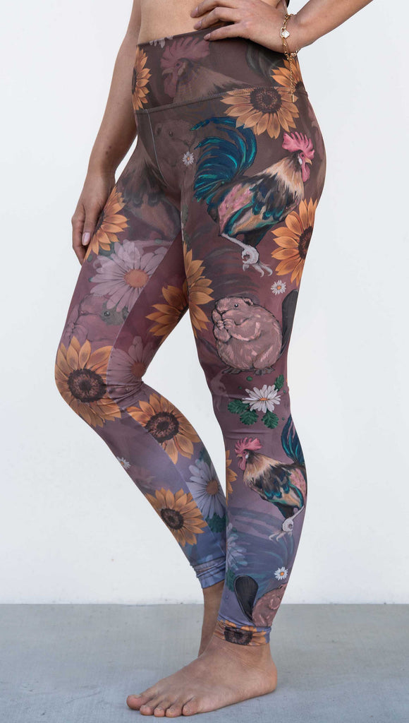 Model wearing WERKSHOP "Cocks n Beavers" Leggings. The leggings show hand painted artwork of roosters and beavers with daisies and sunflowers over an ombreback ground that goes from a warm brown on top to slate blue at the leg opening.