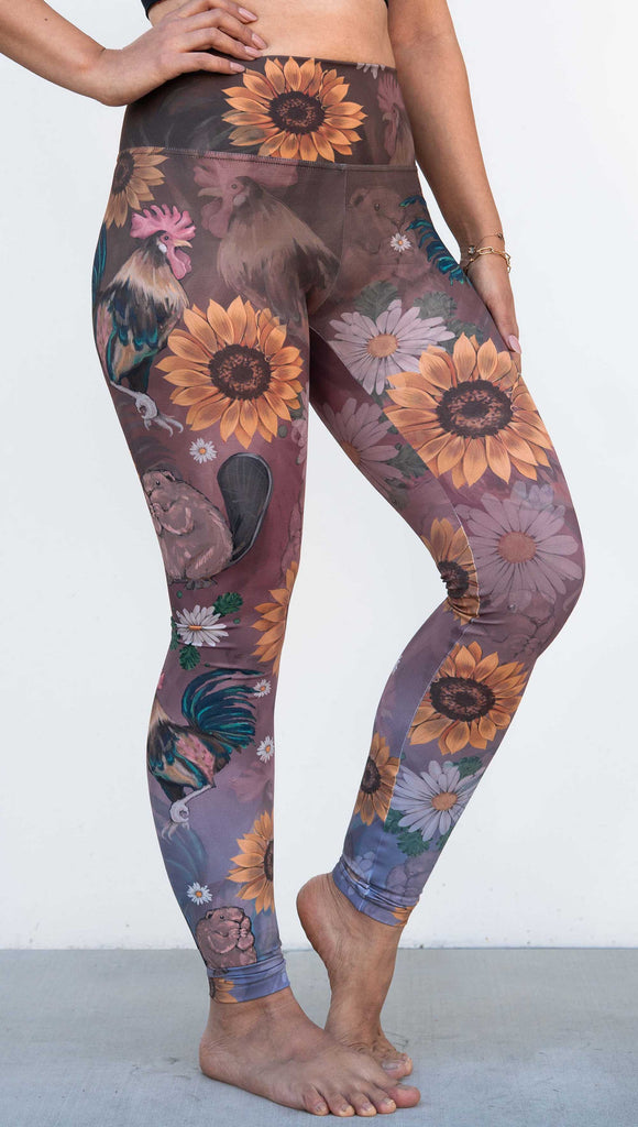 Model wearing WERKSHOP "Cocks n Beavers" Leggings. The leggings show hand painted artwork of roosters and beavers with daisies and sunflowers over an ombreback ground that goes from a warm brown on top to slate blue at the leg opening.
