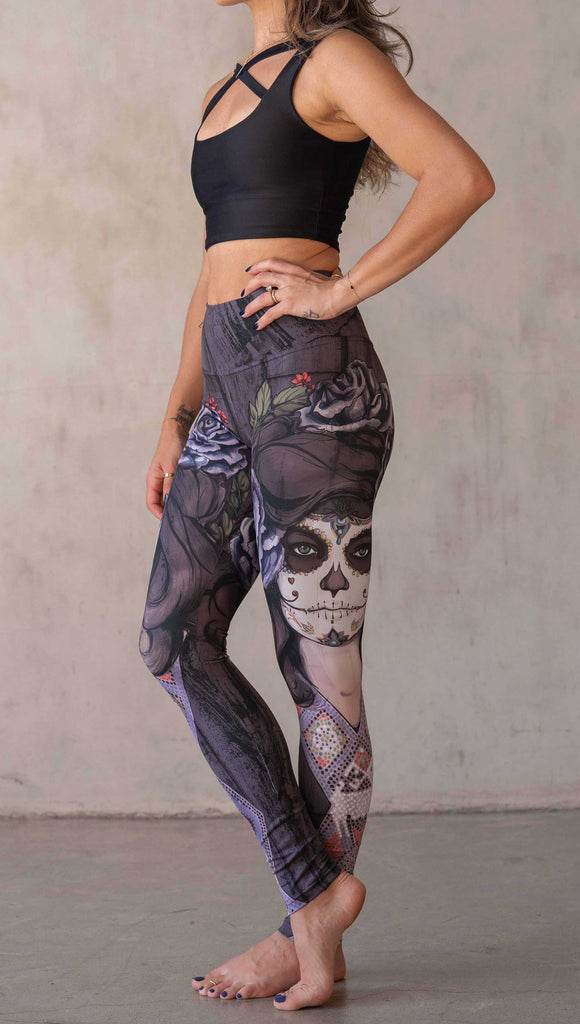 Girl wearing WERKSHOP "Dark Sugar" Athleisure leggings. The artwork on the leggings features a girl wearing dia de los muertos makeup in tones of blue and gray. she is wearing a crown of roses and a beaded necklace over a distressed warm gray woodgrain background.
