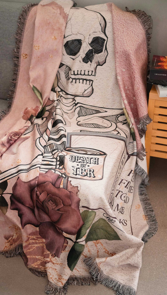 Blanket with a skeleton and floral design on a textured surface. with the phrases, "Death by TBR" and "How to Find Time to Read more Books". (draped over the edge of a futon)