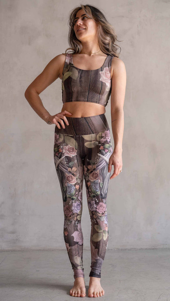 Model wearing WERKSHOP Dark Unicorn Athleisure Leggings. The leggings feature original artwork of a unicorn surrounded by a wreath of vintage coloured roses. The unicorn has a beaded chain with a skull dangling from it's horn. The artwork is over a warm woodgrain background.