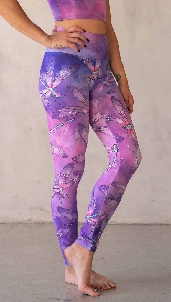 Side view of model wearing WERKSHOP Dragonflies Athleisure leggings. The artwork on the leggings features hand drawn dragonflies with pops of teal and pink over a purple and pink watercolour background.