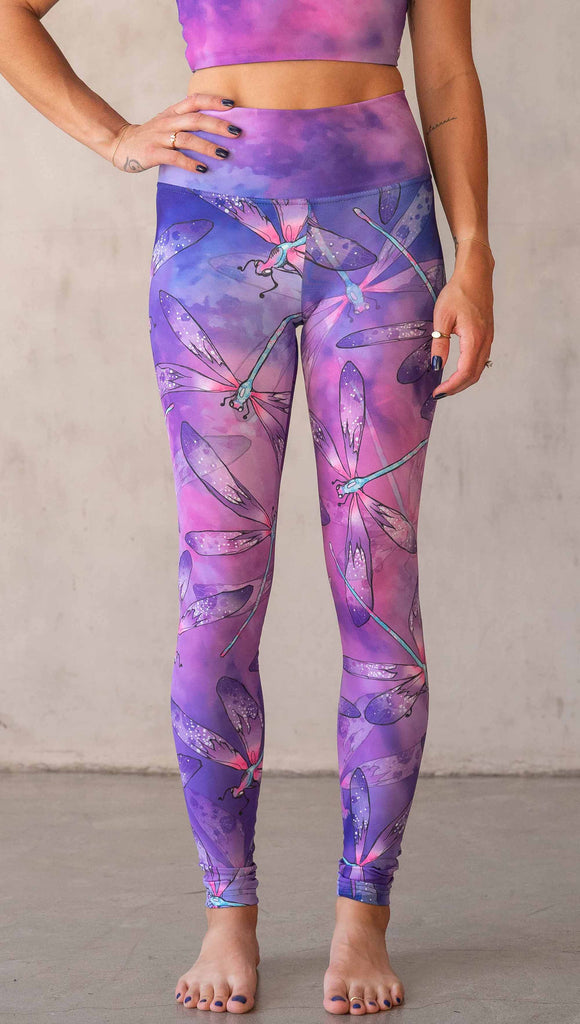 Front view of model wearing WERKSHOP Dragonflies Athleisure leggings. The artwork on the leggings features hand drawn dragonflies with pops of teal and pink over a purple and pink watercolour background.