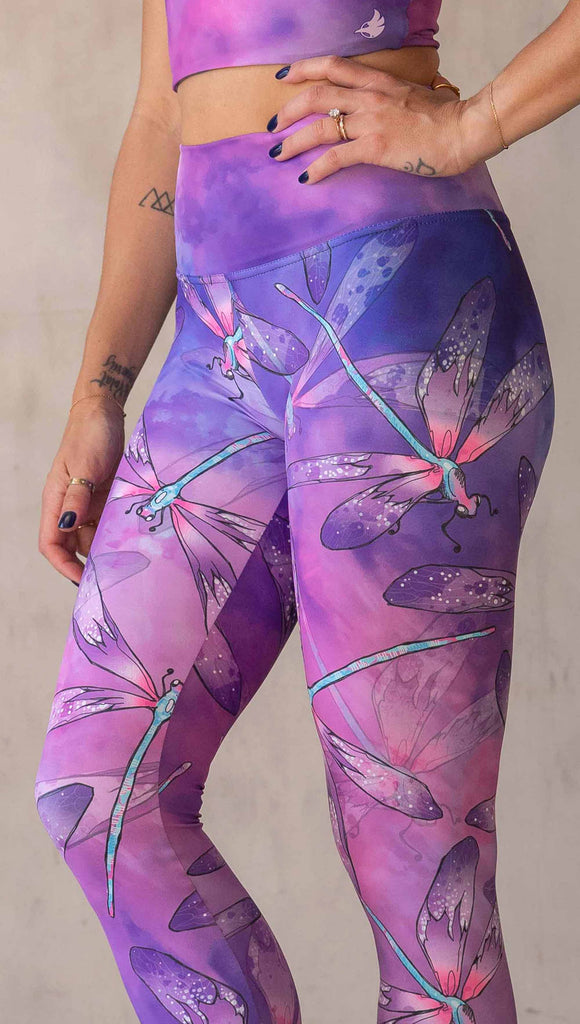Zoomed in view of model wearing WERKSHOP Dragonflies Athleisure leggings. The artwork on the leggings features hand drawn dragonflies with pops of teal and pink over a purple and pink watercolour background.