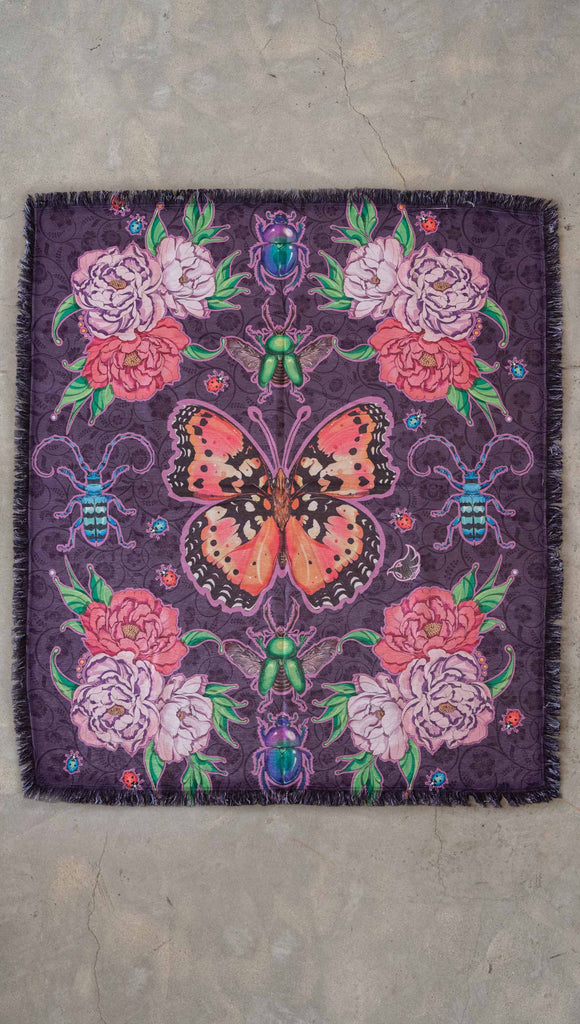 WERKSHOP Enchanted Garden tapestry draped on a couch. The tapestry is printed with original artwork by Chriztina Marie. The artwork printed not the fabric features Butterflies, Beetles and Peonies over a warm fuchsia with bright bold pops of color on each beetle and Butterfly.