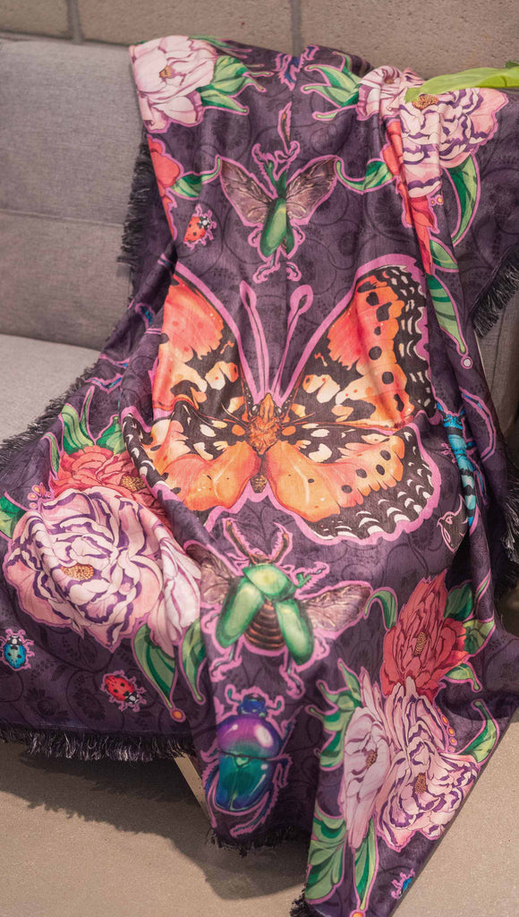 WERKSHOP Enchanted Garden tapestry draped on a couch. The tapestry is printed with original artwork by Chriztina Marie. The artwork printed not the fabric features Butterflies, Beetles and Peonies over a warm fuchsia with bright bold pops of color on each beetle and Butterfly.