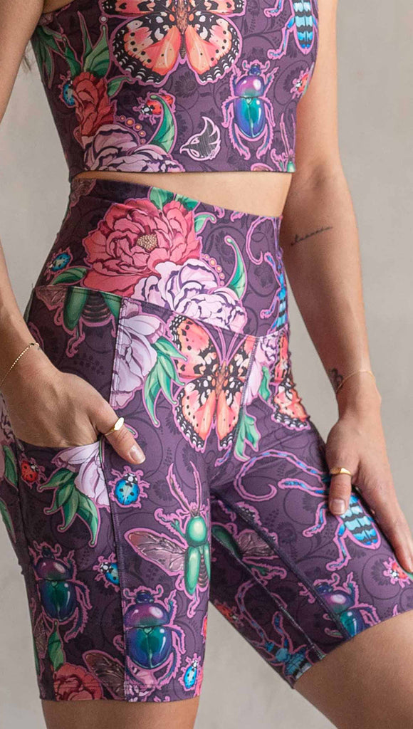 Zoomed in view of model wearing WERKSHOP Enchanted Garden EnviSoft Bicycle length shorts with pockets. The fabric is printed with original artwork by our Female Founder, Chriztina Marie. The artwork printed not the fabric features Butterflies, Beetles and Peonies over a warm fuchsia with bright bold pops of color on each beetle and Butterfly.