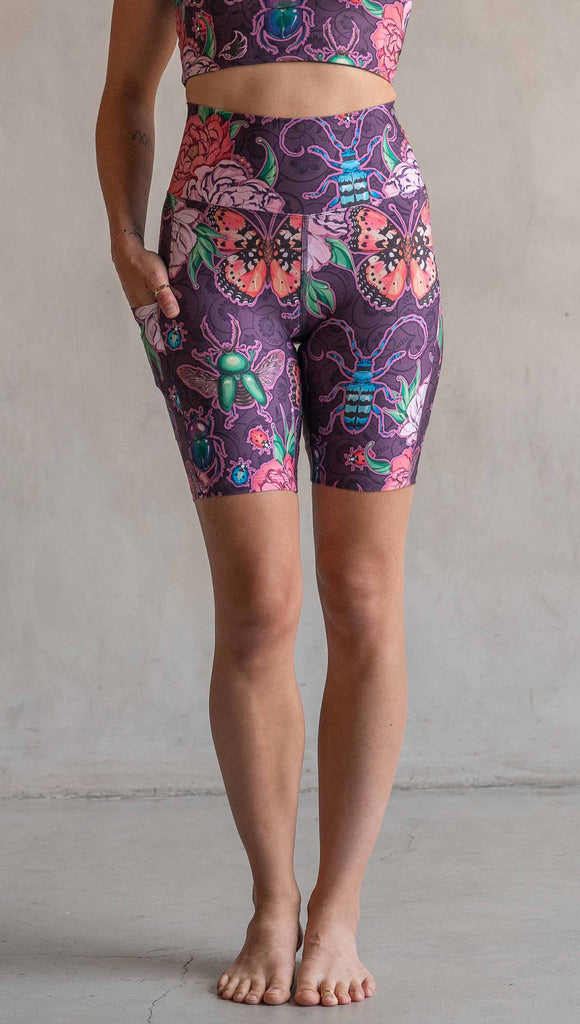 Front view of model wearing WERKSHOP Enchanted Garden EnviSoft Bicycle length shorts with pockets. The fabric is printed with original artwork by our Female Founder, Chriztina Marie. The artwork printed not the fabric features Butterflies, Beetles and Peonies over a warm fuchsia with bright bold pops of color on each beetle and Butterfly.