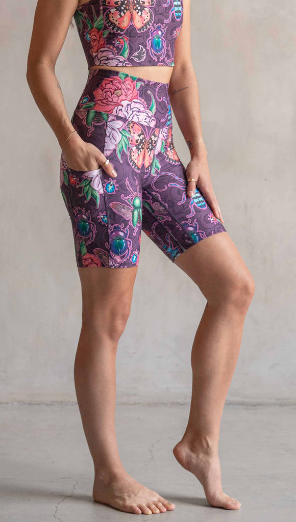 Side view of model wearing WERKSHOP Enchanted Garden EnviSoft Bicycle length shorts with pockets. The fabric is printed with original artwork by our Female Founder, Chriztina Marie. The artwork printed not the fabric features Butterflies, Beetles and Peonies over a warm fuchsia with bright bold pops of color on each beetle and Butterfly.