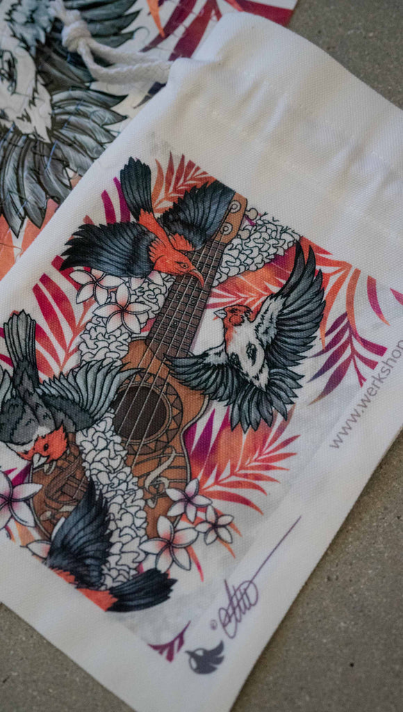 WERKSHOP Ukulele jigsaw puzzle. The artwork features a flower lei wrapped around a ukulele with red and black bird flying. The background has coral and purple watercolor palms. The puzzle comes with a canvas drawcord pouch.