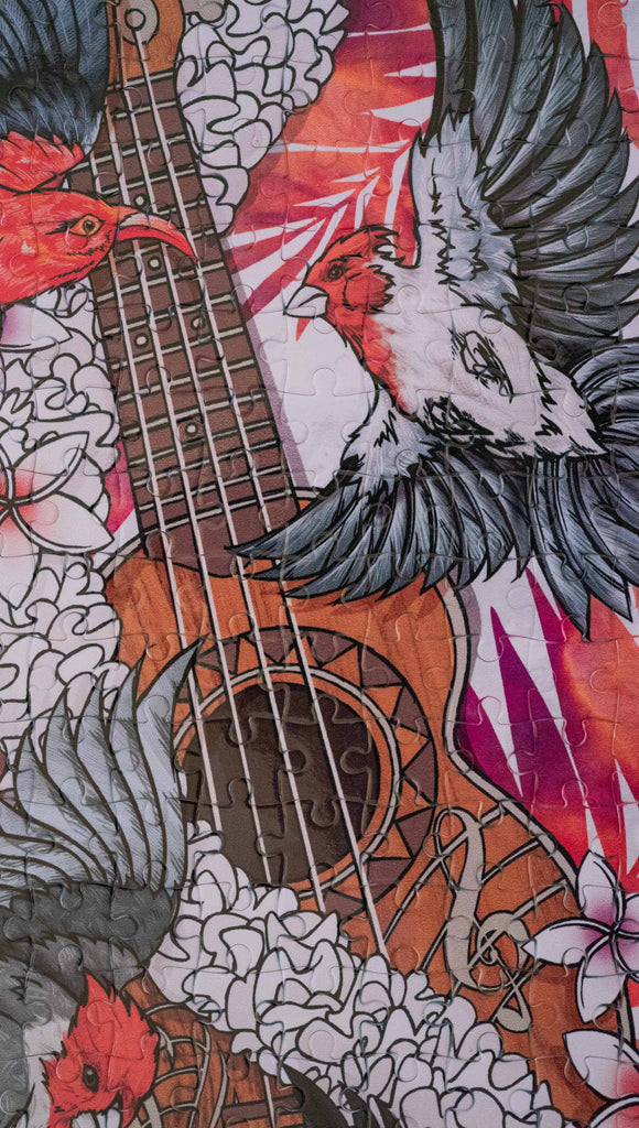 WERKSHOP Ukulele jigsaw puzzle. The artwork features a flower lei wrapped around a ukulele with red and black bird flying. The background has coral and purple watercolor palms. The puzzle comes with a canvas drawcord pouch.