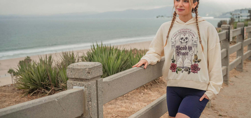 Person wearing a hoodie with a graphic design by a beach. The bookish hoodie features a tombstone with skull and flowers and an open book. On the tombstone, it says: "Famous Last Words: Just One More Chapter" and "Read in Peace"
