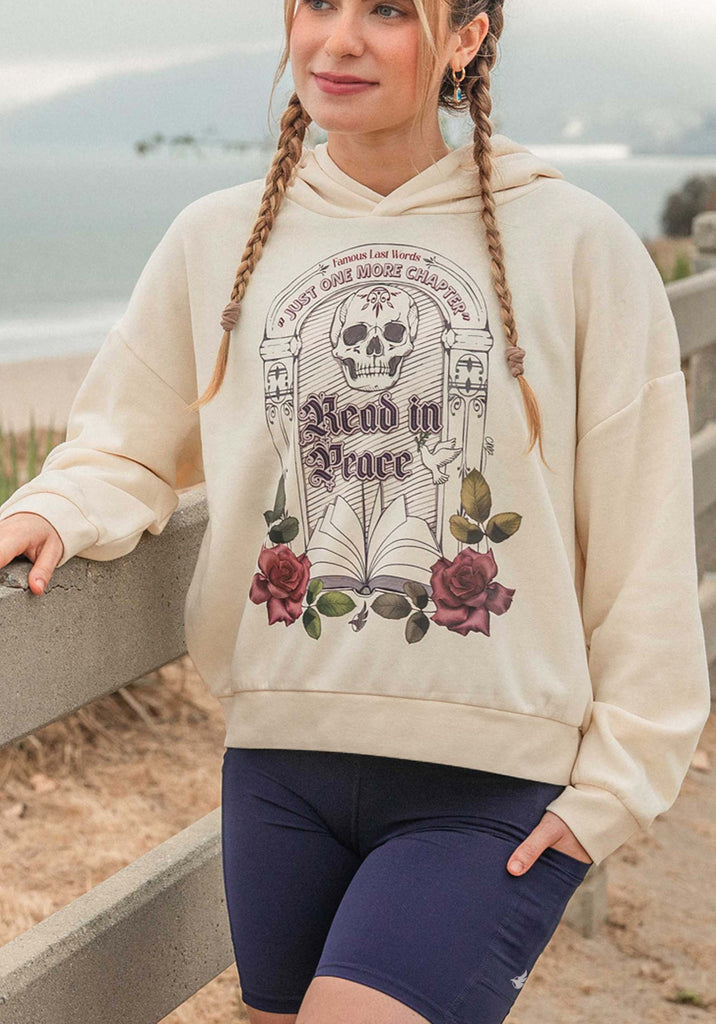 Person wearing a hoodie with a graphic design by a beach. The bookish hoodie features a tombstone with skull and flowers and an open book. On the tombstone, it says: "Famous Last Words: Just One More Chapter" and "Read in Peace"