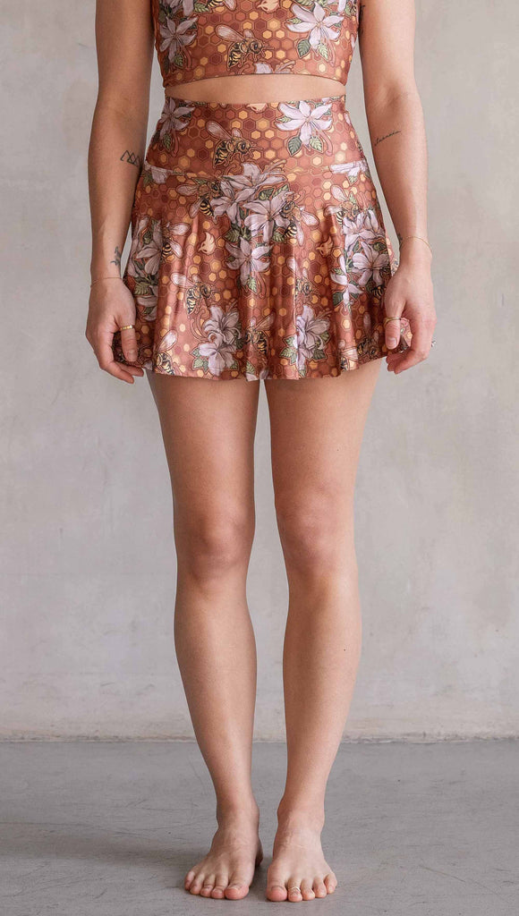 Model wearing WERKSHOP Featherlight set with Honeybess Skirt and Top. The skirt features built-in shorts with pockets. The fabric is printed with original artwork by Chriztina Marie that features clusters of honeybees and flowers over a gold honeycomb inspired background. The skirt falls to about the wearer's fingertips and the shorts are hidden underneath.