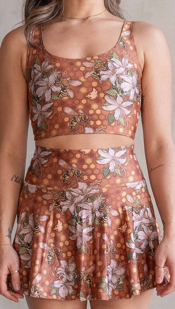 Zoomed in photo of model wearing WERKSHOP Featherlight set with Honeybess Skirt and Top. The skirt features built-in shorts with pockets. The fabric is printed with original artwork by Chriztina Marie that features clusters of honeybees and flowers over a gold honeycomb inspired background. The skirt falls to about the wearer's fingertips and the shorts are hidden underneath.