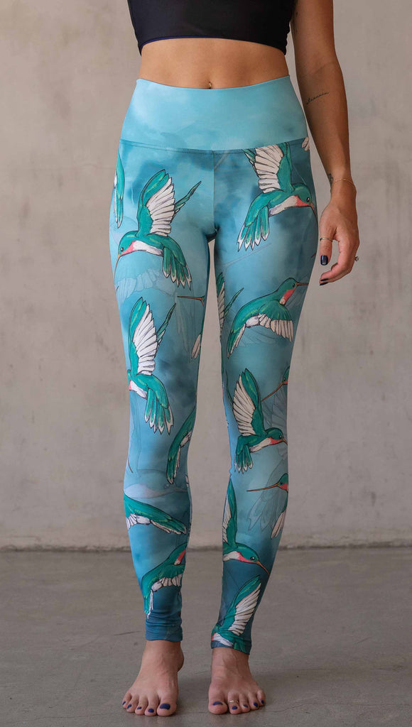 Front view of model wearing WERKSHOP Hummingbirds athleisure leggings. The artwork on the leggings fetures hummingbirds fluttering through the sky over an aqua watercolor background.