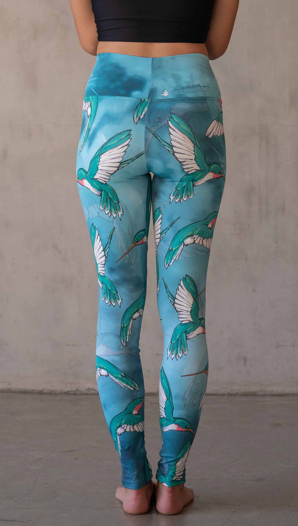 Back view of model wearing WERKSHOP Hummingbirds athleisure leggings. The artwork on the leggings fetures hummingbirds fluttering through the sky over an aqua watercolor background