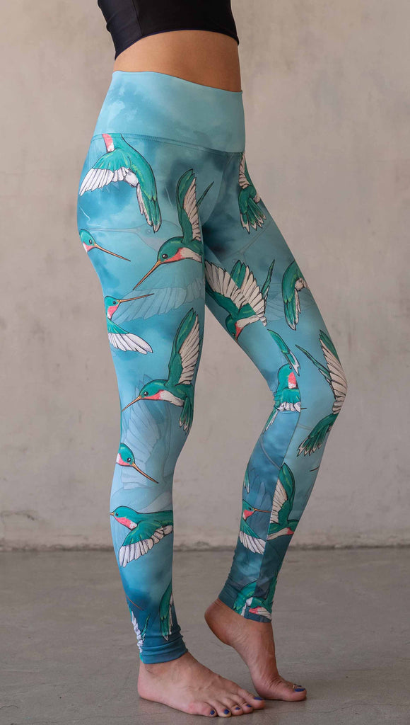 Side view of model wearing WERKSHOP Hummingbirds athleisure leggings. The artwork on the leggings fetures hummingbirds fluttering through the sky over an aqua watercolor background