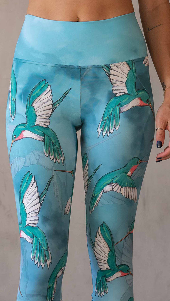 Zoomed in view of model wearing WERKSHOP Hummingbirds athleisure leggings. The artwork on the leggings fetures hummingbirds fluttering through the sky over an aqua watercolor background