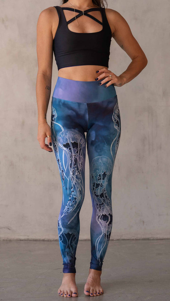 Model wearing WERKSHOP Jellyfish Athleisure Leggings. The artwork on the leggings shows white jellyfish swimming up the wearer's legs over a cobalt blue and purple watercolor background.
