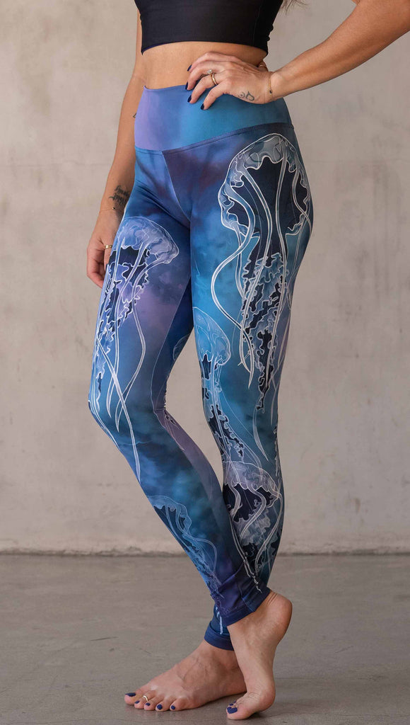 Model wearing WERKSHOP Jellyfish Athleisure Leggings. The artwork on the leggings shows white jellyfish swimming up the wearer's legs over a cobalt blue and purple watercolor background.
