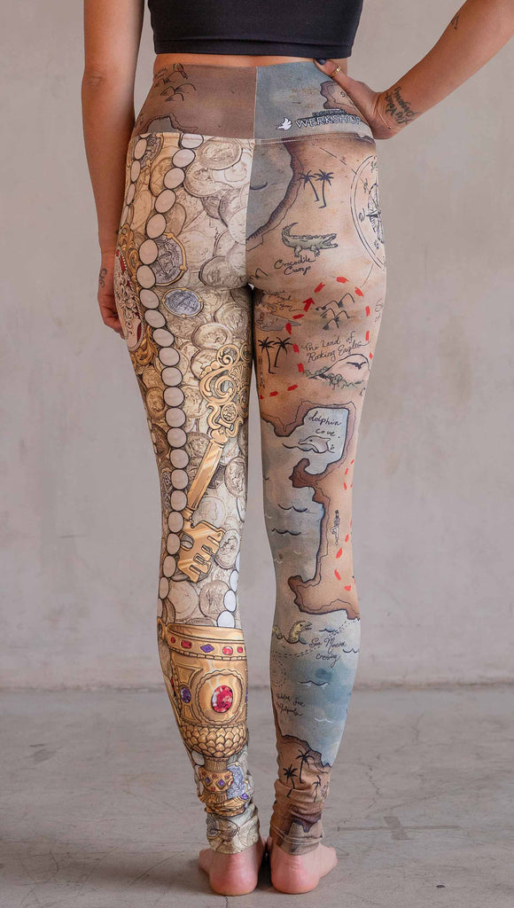 Model wearing WERKSHOP Booty Mashup Leggings. The wearer's right leg is printed with vintage inspired, hand drawn, treasure map artwork and the left leg is golden treasure with a kings royal crown, keys and pearls: everything you'd expect to find inside a pirates treasure.