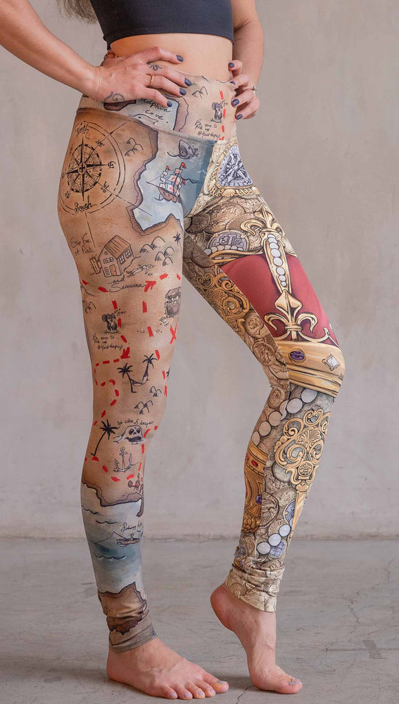 Model wearing WERKSHOP Booty Mashup Leggings. The wearer's right leg is printed with vintage inspired, hand drawn, treasure map artwork and the left leg is golden treasure with a kings royal crown, keys and pearls: everything you'd expect to find inside a pirates treasure.