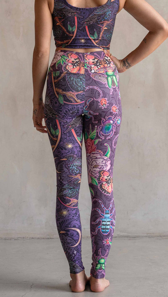 Back view of model wearing WERKSHOP Starlings and Enchanted Garden Mashup Leggings. The fabric is printed with original artwork by Chriztina Marie. Each leg features different artwork. The wearer’s left leg has European Starlings perched on a branch near a crescent moon and fireflies. The colors are warm purples with pops of pink, gold and green. The wearer’s right leg features Butterflies, Beetles and Peonies over a warm fuchsia with bright bold pops of color on each beetle and Butterly.