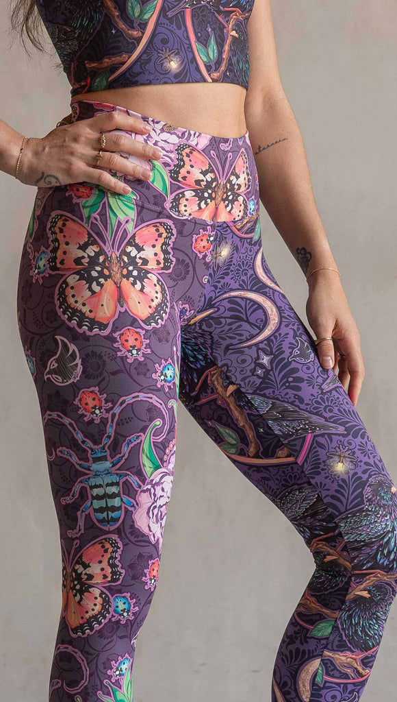 Zoomed in view of model wearing WERKSHOP Starlings and Enchanted Garden Mashup Leggings. The fabric is printed with original artwork by Chriztina Marie. Each leg features different artwork. The wearer’s left leg has European Starlings perched on a branch near a crescent moon and fireflies. The colors are warm purples with pops of pink, gold and green. The wearer’s right leg features Butterflies, Beetles and Peonies over a warm fuchsia with bright bold pops of color on each beetle and Butterly.