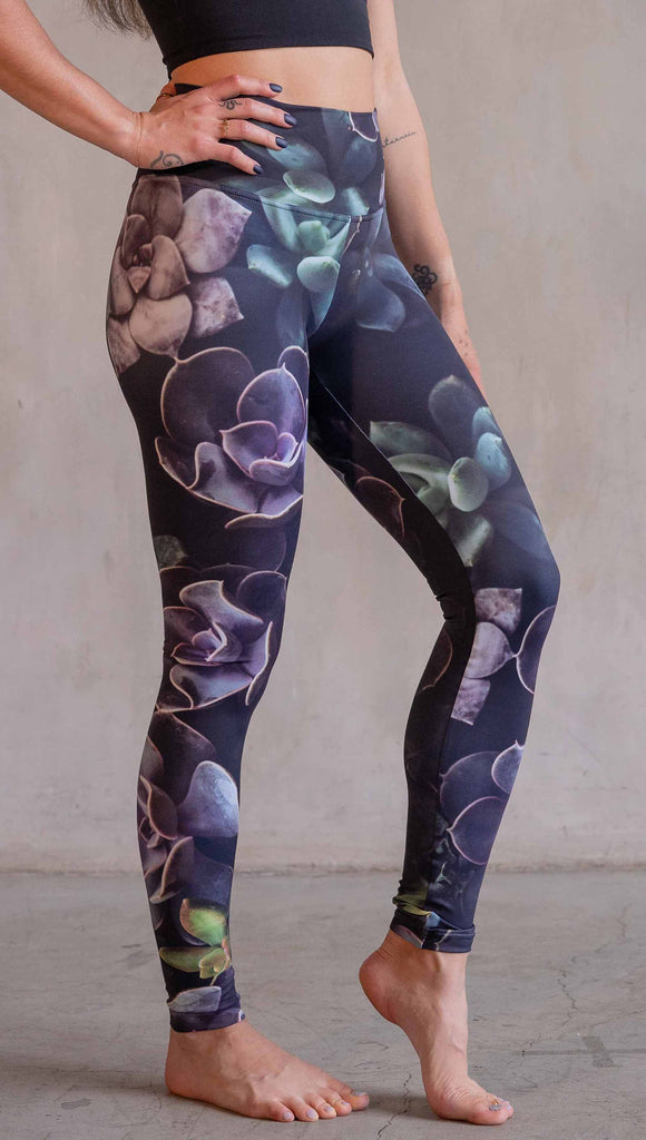 Model wearing WERKSHOP Midnight Garden Athleisure Leggings. The artwork on the leggings is printed with succulents in dark romantic colors of purple with small pops of teal