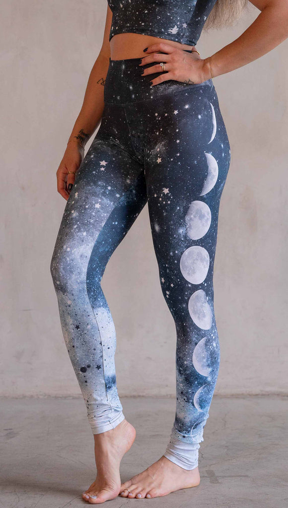 Model wearing WERKSHOP Moon Phases Athleisure Leggings. The leggings feature the phases of the moon down the wearer's left leg over a starry blue galactic background. All 12 zodiac constellations are sprinkled throughout the background of the design.