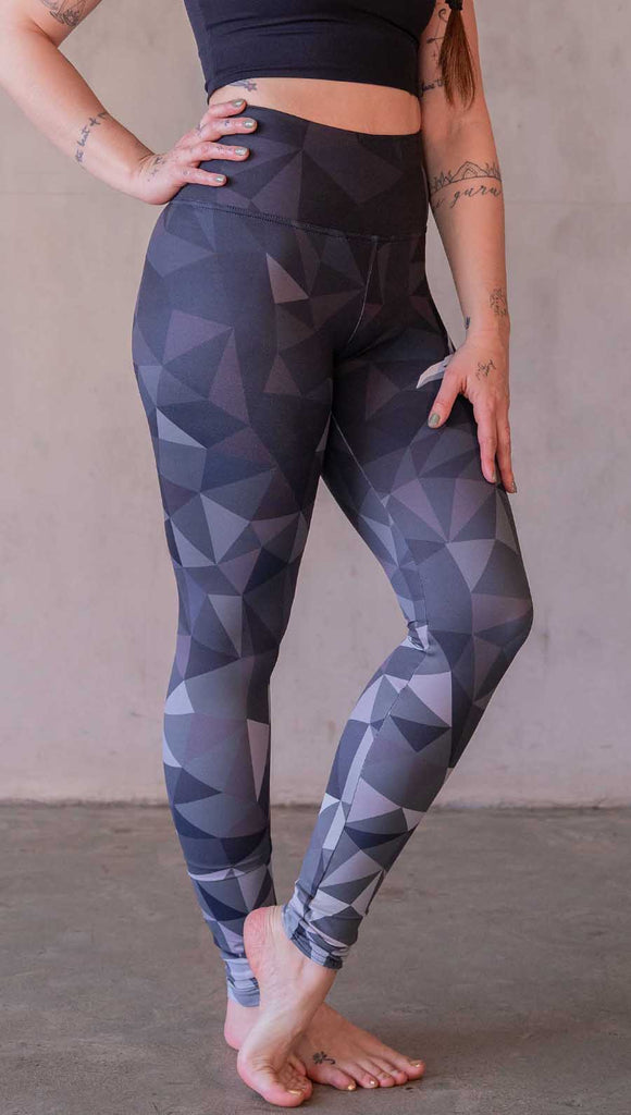 Model wearing WERKSHOP Onyx leggings featuring original and exclusive polygon artwork that has a dark gray and black at the waistband to light gray ombre fade below the knee. The design is asymmetrical with the WERKSHOP eagle logo is on the wearers left hip. 