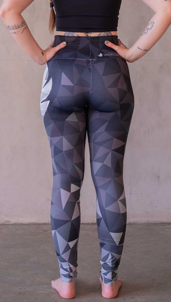 Model wearing WERKSHOP Onyx leggings featuring original and exclusive polygon artwork that has a dark gray and black at the waistband to light gray ombre fade below the knee. The design is asymmetrical with the WERKSHOP eagle logo is on the wearers left hip. 