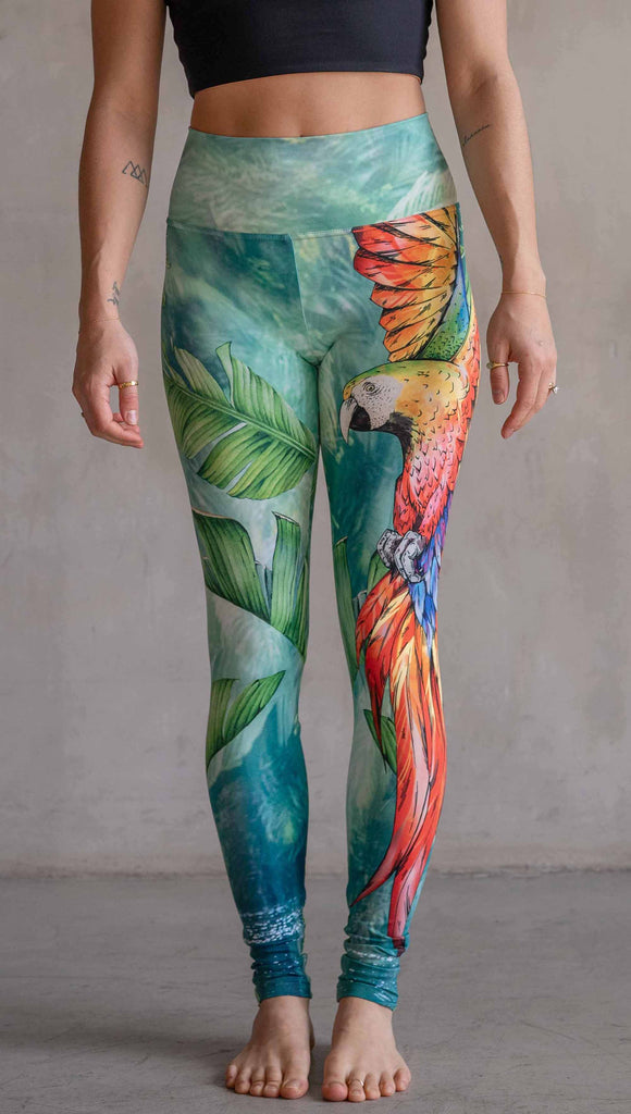Model wearing WERKSHOP Parrot Athleisure Leggings. The artwork on the leggings feature a tropical parrot down the wearer's left leg and bright green and teal floral artwork on the right leg.