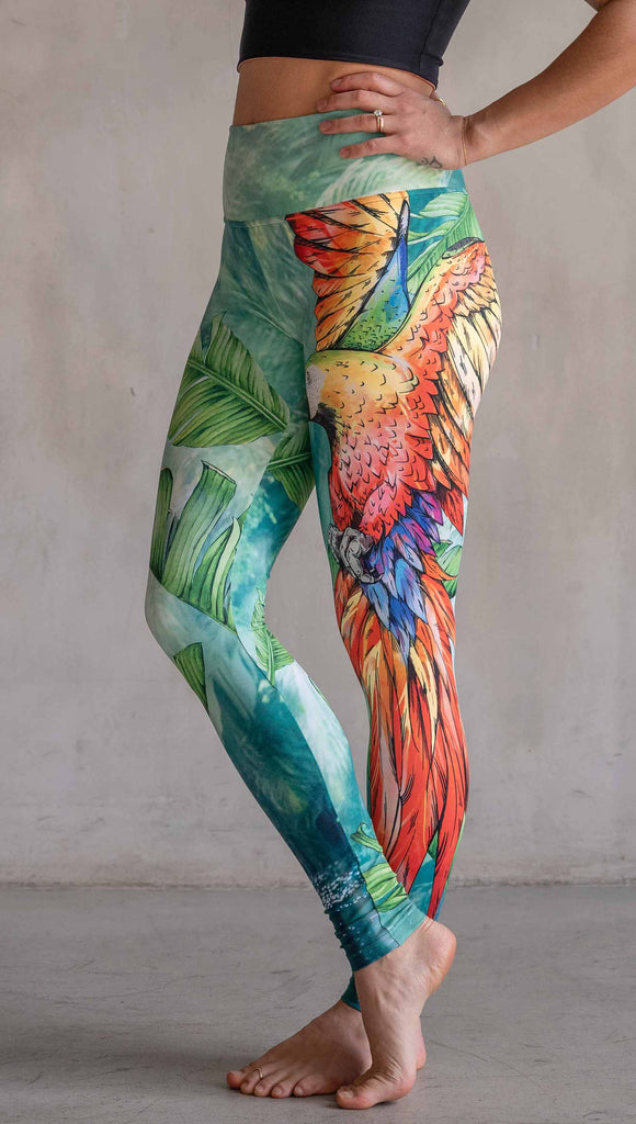 Model wearing WERKSHOP Parrot Athleisure Leggings. The artwork on the leggings feature a tropical parrot down the wearer's left leg and bright green and teal floral artwork on the right leg.