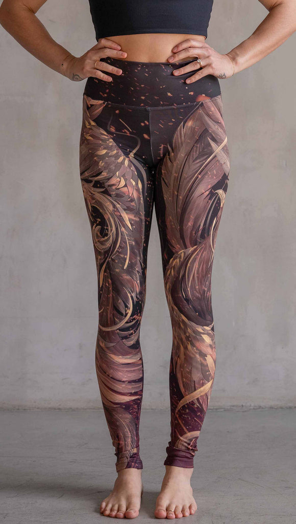 Model wearing WERKSHOP Phoenix Athleisure Leggings. The leggings are printed with a mythical phoenix swirling up each leg with embers flying through the background. The overall color theme is super earthy and warm browns, oranges and yellow tones