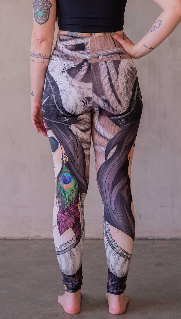 Model wearing WERKSHOP Pirate Girl Athliesure Leggings. The artwork on the leggings feature a sexy pirate with an eye patch, red bandana, hat with skull and cross-bones, and a peacock feather earring. The background is woodgrain and ropes.