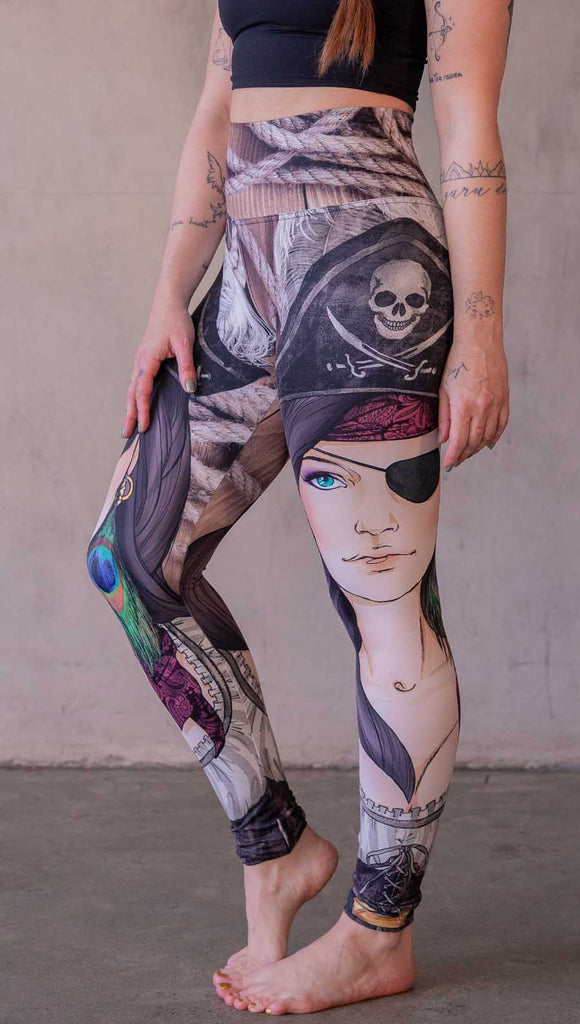 Model wearing WERKSHOP Pirate Girl Athliesure Leggings. The artwork on the leggings feature a sexy pirate with an eye patch, red bandana, hat with skull and cross-bones, and a peacock feather earring. The background is woodgrain and ropes.