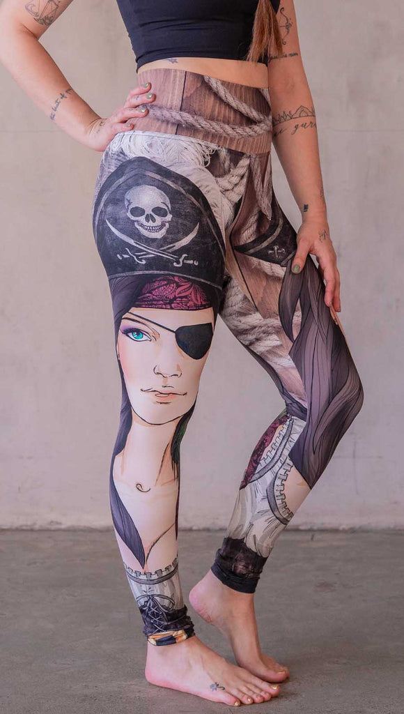 Model wearing WERKSHOP Pirate Girl Athliesure Leggings. The artwork on the leggings feature a sexy pirate with an eye patch, red bandana, hat with skull and cross-bones, and a peacock feather earring. The background is woodgrain and ropes.