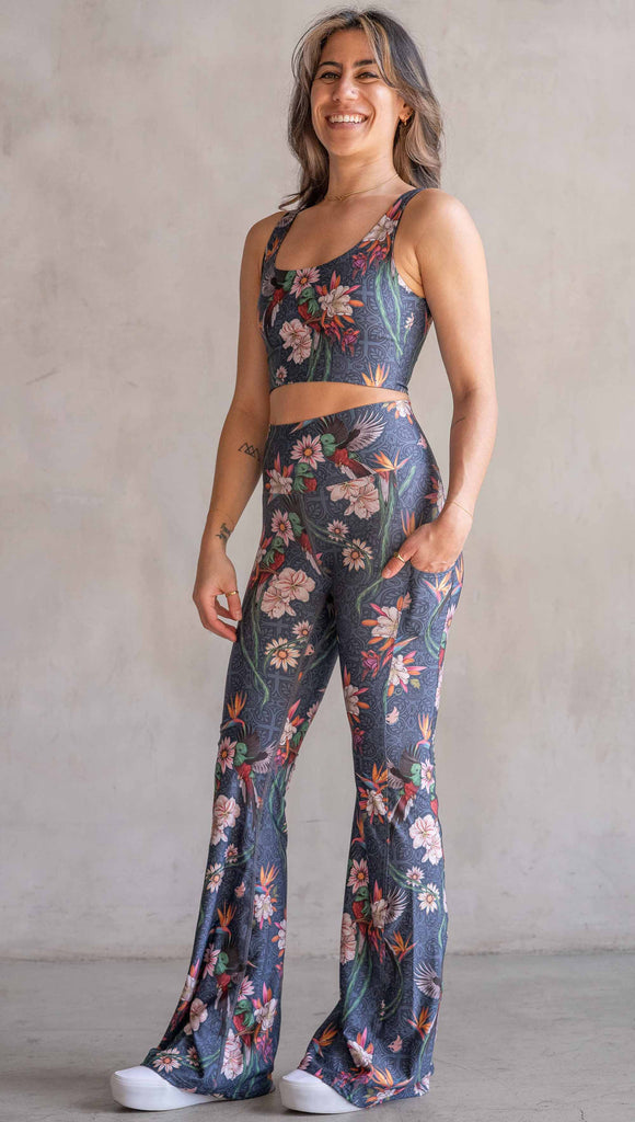 Model wearing WERKSHOP Quetzal Featherlight Bells. The leggings are printed with with original quetzal artwork with clusters of tropical flowers and birds of paradise over a blue background. The featherlight bells have pockets.