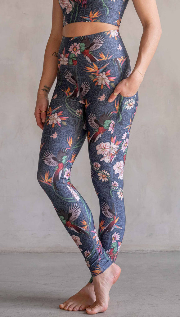 Model wearing WERKSHOP Quetzal Featherlight Leggings. The leggings are printed with with original quetzal artwork with clusters of tropical flowers and birds of paradise over a blue background. The featherlight leggings have pockets.