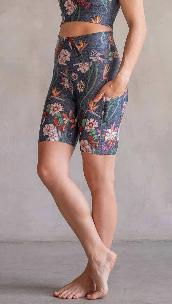 Model wearing WERKSHOP Quetzal Featherlight Bicycle length shorts. The shorts are printed with with original quetzal artwork with clusters of tropical flowers and birds of paradise over a blue background. The featherlight shirt have pockets.