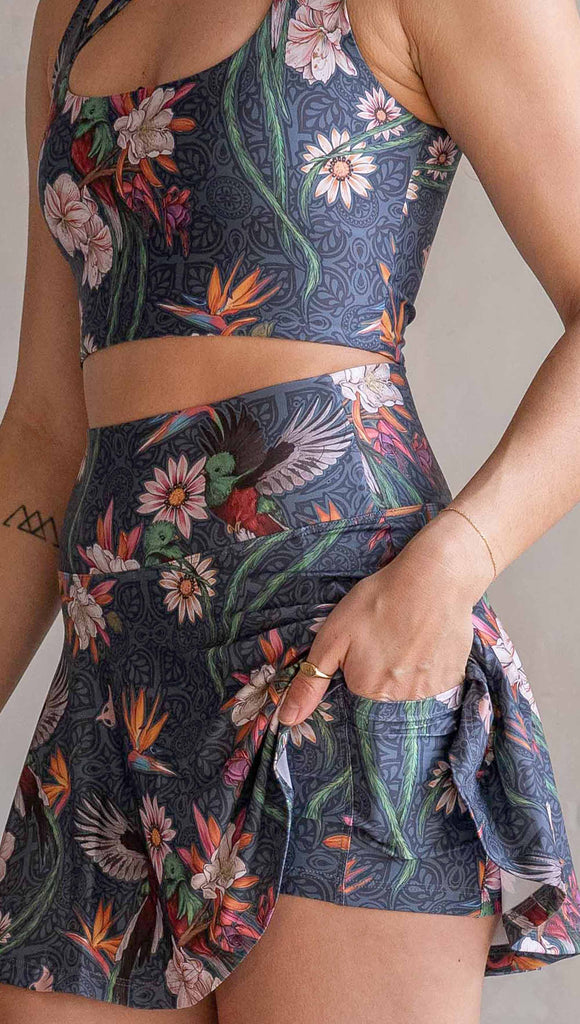 Model wearing WERKSHOP Quetzal Featherlight Active Skirt. The skirts have built in shorts underneath and are printed with with original quetzal artwork with clusters of tropical flowers and birds of paradise over a blue background. The featherlight skirts have pockets.