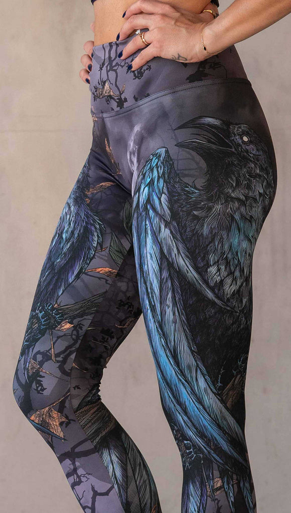 Zoomed in image of girl wearing WERKSHOP "Raven" athleisure leggings. The artwork on the leggings feature a raven perched on a branch looking into the moonlight with pops of blue glistening from his feathers. He is framed by autumn leaves over a very spooky night sky.