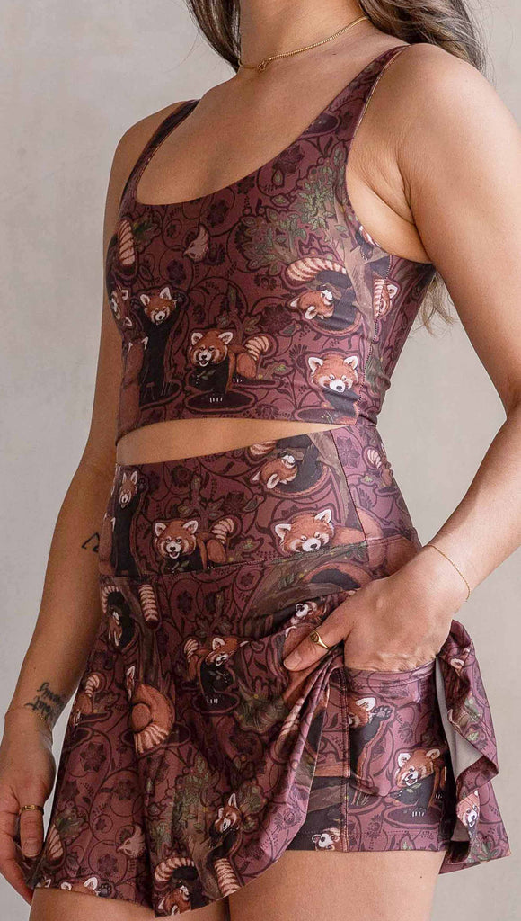 Zoomed in view of model wearing WERKSHOP Featherlight set with Red Panda skirt and matching top. The skirt features built-in shorts underneath with a pocket. The artwork printed on the fabric is the original/personal artwork of Chriztina Marie. It has clusters of tasteful and adorable red pandas playing on trees over a burgundy background. The skirt is flirty with a length that goes to about the wearer's fingertips.