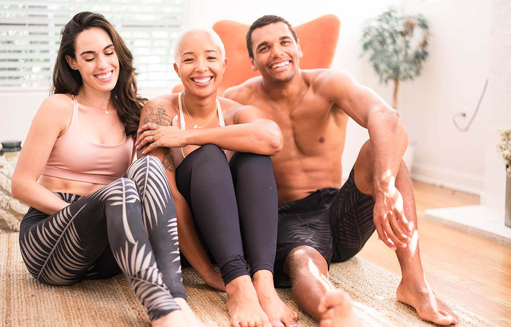 3 friends casually sitting on the floor and laughing together. The 1st girl is wearing black palms leggings, the 2nd girl is wearing solid black leggings and the guy is wearing black palms performance shorts.