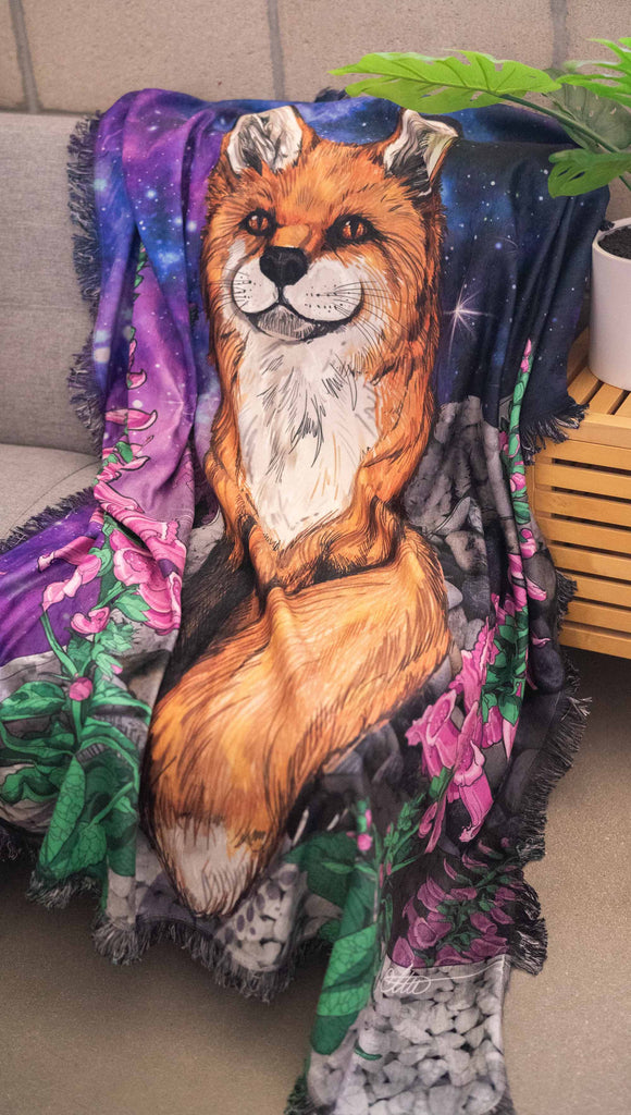 WERKSHOPs Save A Fox Tapestry draped on a futon. The tapestry features soft chenille fabric, with original artwork of a fox in front of a galactic background with foxglove plants framing him.