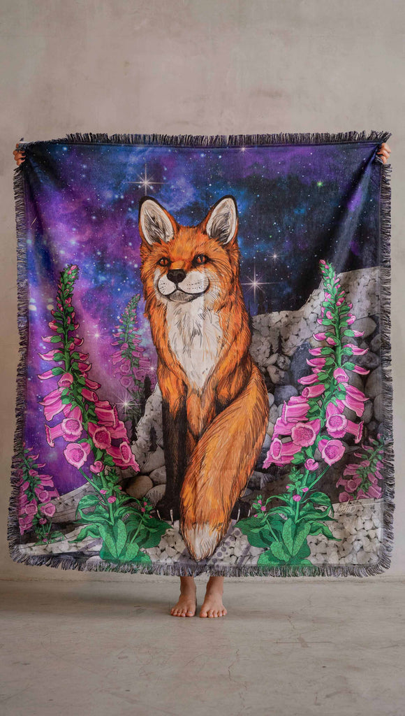 WERKSHOPs Save A Fox Tapestry draped on a futon. The tapestry features soft chenille fabric, with original artwork of a fox in front of a galactic background with foxglove plants framing him.