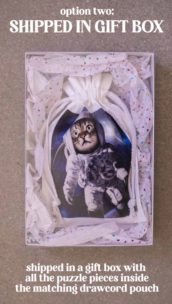 WERKSHOP Space Cat (Catstronaut) Puzzle. The artwork features a house cat wearing an astronaut uniform, floating in outer space with a nebula behind him. Option 2: shipped in a gift box with all the puzzle pieces inside the matching drawcord pouch.